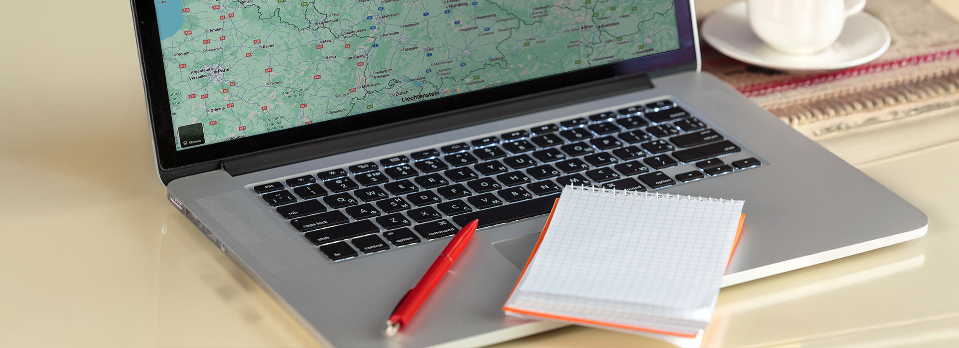 Planung im Büro bei Theater Liberi Laptop mit geöffneter Landkarte, daneben ein roter Stift, ein kariertes Notizbuch und ein Kaffee – Symbol für die Arbeit im Büro bei Theater Liberi.