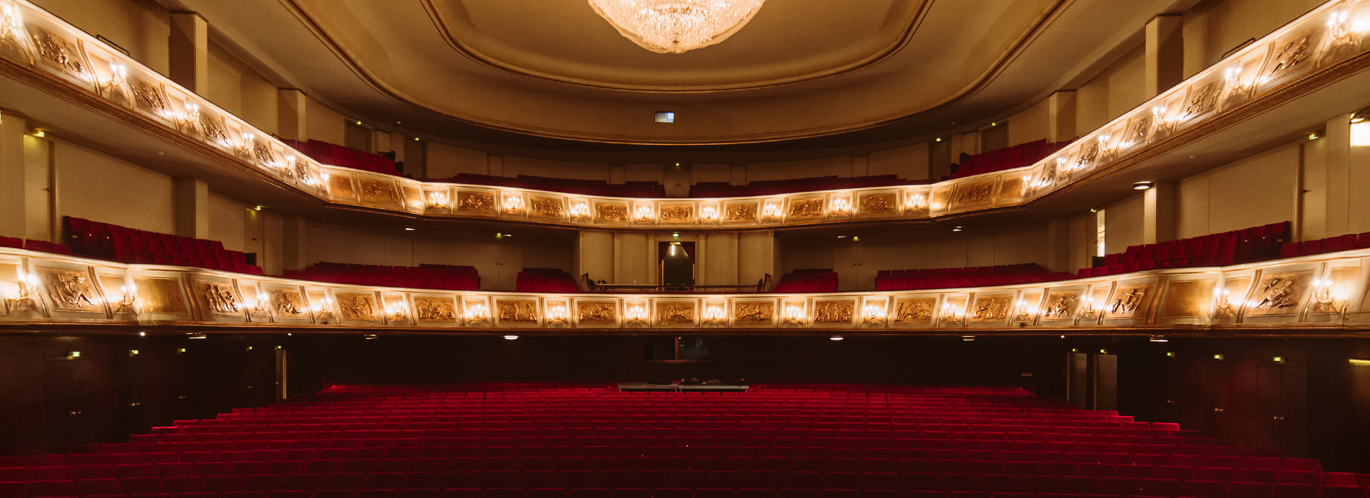 theater-liberi-theatersaal-vorstellung-tickets.jpg Blick aus dem Zuschauerraum auf die leeren roten Sitzreihen eines prunkvollen Theatersaals mit drei Rängen und einem großen Kronleuchter.