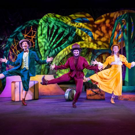 theater-liberi-musical-tanzszene-farbenfroh-peter-harbauer Drei Darsteller in bunten Kostümen tanzen vor lebendigem Bühnenbild.