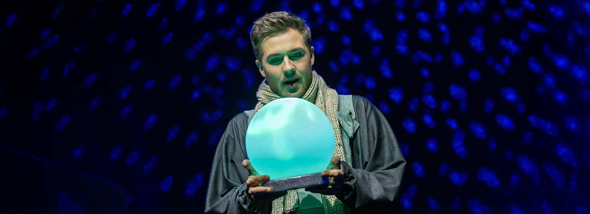 Schauspieler auf der Bühne hält eine leuchtende, türkisfarbene Kristallkugel, während er in einem dramatisch beleuchteten Bühnenbild mit blauem Sternenlicht steht.