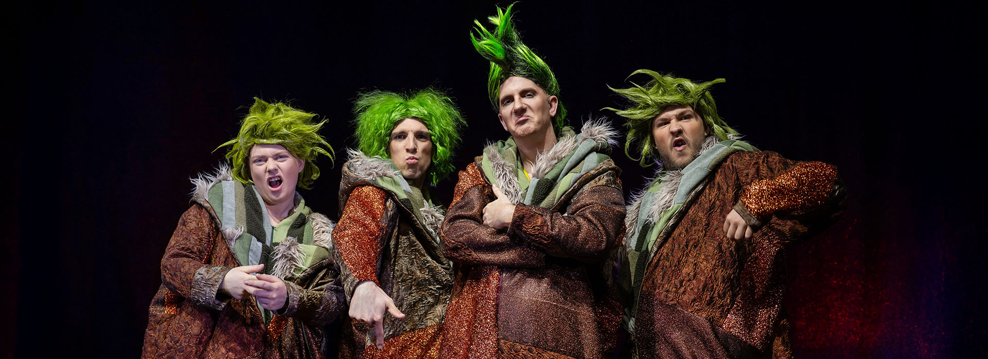 Die frechen Schlawuzis aus „Schneekönigin – das Musical“ Vier Darsteller in glitzernden, braun-orangenen Schlawuzi-Kostümen mit wilden, grünen Perücken stehen auf der Bühne und machen freche Posen.