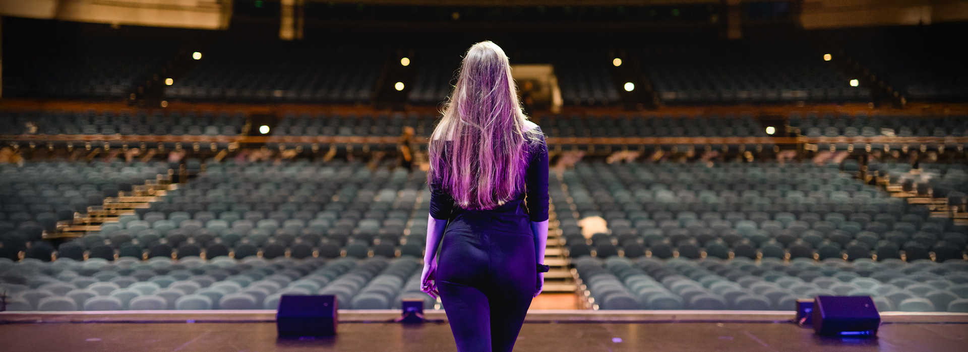 Darstellerin vor leerem Saal Eine Frau mit langen Haaren steht auf einer Theaterbühne und blickt in einen leeren, großen Zuschauerraum mit hunderten blauen Sitzen.