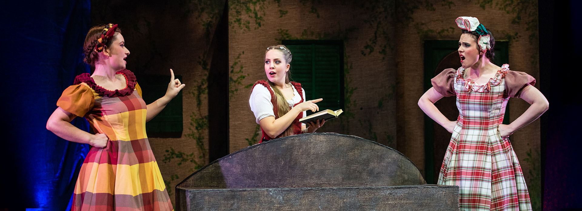 Belle mit ihren Schwestern in „Die Schöne und das Biest – das Musical“ Drei Darstellerinnen in bunten Kostümen stehen um einen großen Brunnen; eine von ihnen hält ein Buch – es ist Belle.