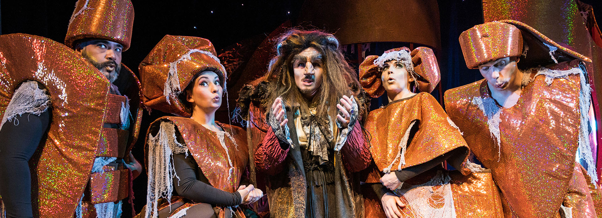 Das Biest mit verzauberten Dienern aus „Die Schöne und das Biest – das Musical“ Eine Gruppe Darsteller in fantasievollen, kupferfarbenen Kostümen steht um das Biest mit zotteliger Mähne in der Mitte.