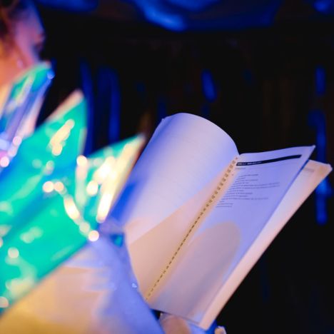 theater-liberi-backstage-skript-einblick-inszenierung.jpg Eine Person hält ein aufgeschlagenes Skript in der Hand, während sie in einem Kostüm mit blaugrünen Spitzen auf den Auftritt wartet.