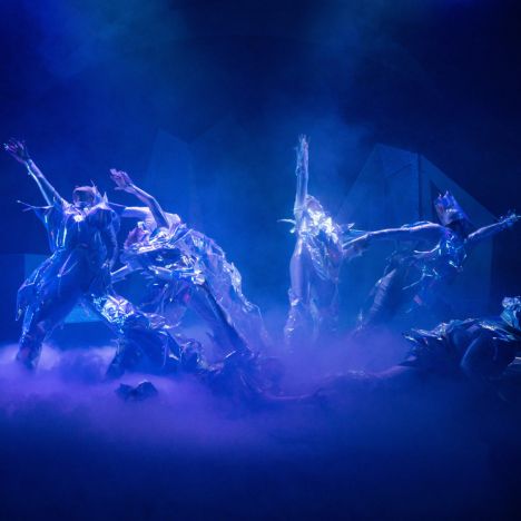 theater-liberi-tanzszene-buehnennebel-showmoment.jpg Tänzer:innen in reflektierenden silber-blauen Kostümen bewegen sich dramatisch im Bühnennebel.