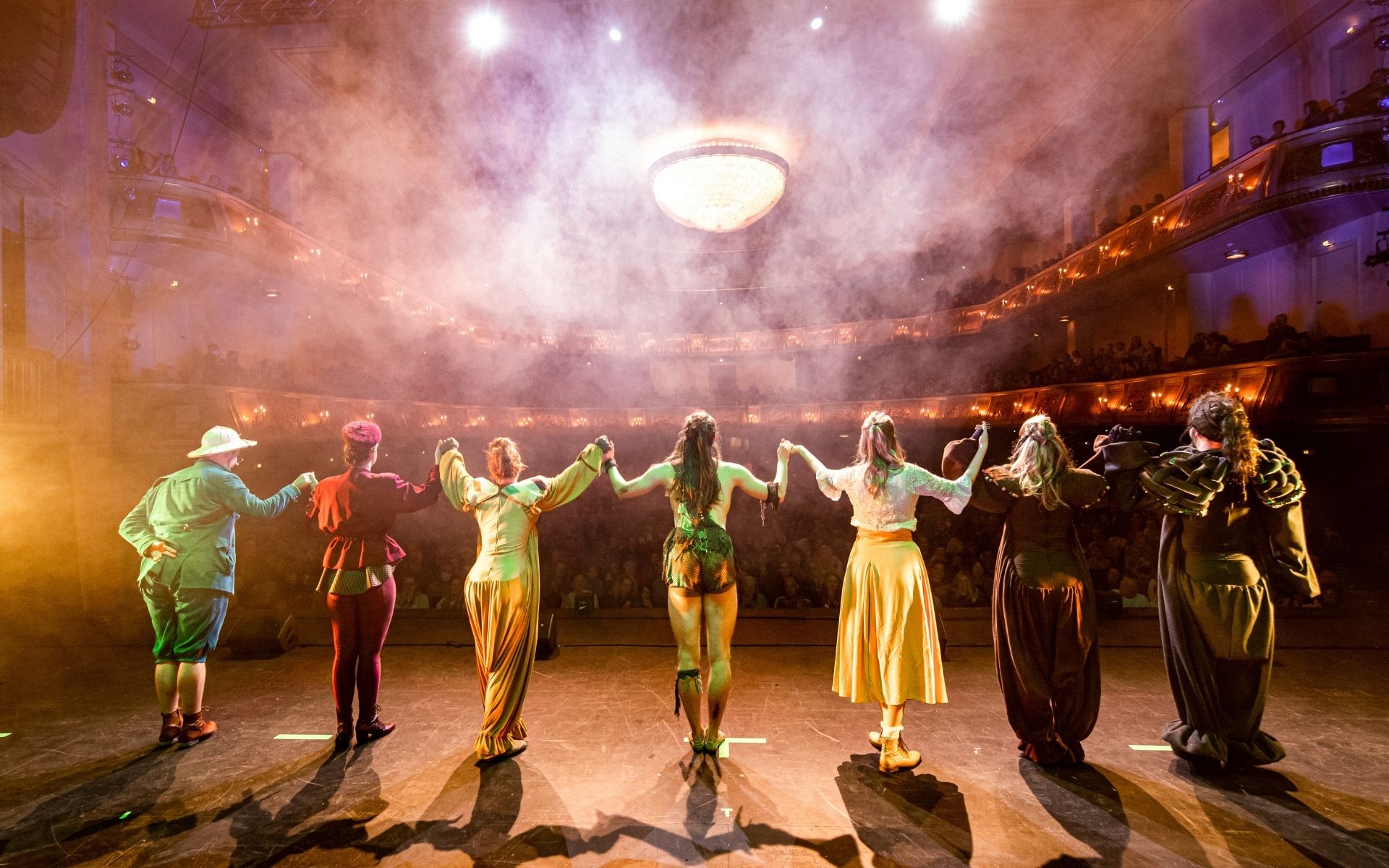 theater-liberi-vorhangschluss-applaus-buehne.jpg Ensemble steht Hand in Hand auf der Bühne vor Publikum im Theaterlicht.